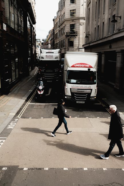 A street scene in an urban area with narrow roads and tall, multi-story buildings on both sides during daytime. Two delivery trucks, one with 'Uniserve' branding and the other with an unspecified logo, are parked along the curb, partially extending into the road to facilitate loading or unloading. A black motorcycle is parked next to the trucks on the pavement. In the foreground, a woman dressed in dark clothing carrying a black bag is crossing the street, followed by an older man with white hair wearing a dark coat and lighter pants also walking across the crosswalk. The environment appears to be related to home relocation or moving services, with the vehicles used for furniture transport or packing activities by [COMPANY_NAME], set in a typical cityscape that emphasizes the logistical aspect of moving in an urban setting.