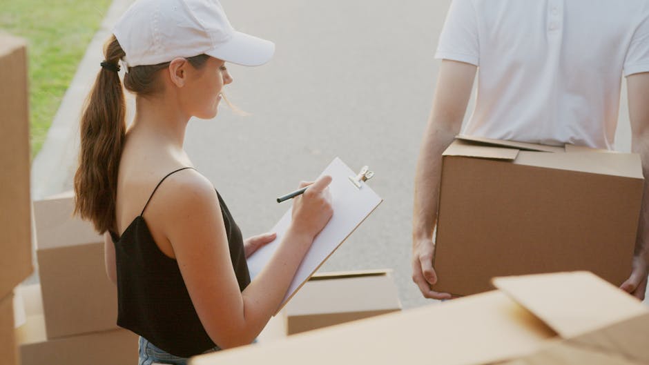 A woman wearing a white cap and a black spaghetti strap top is outdoors, holding a clipboard and writing with a pen. In front of her, a person in a white t-shirt is carrying a large, sealed cardboard box towards a collection of other moving boxes placed on the pavement. The scene appears to be part of a home relocation process, with several cardboard boxes of various sizes wrapped in plastic or filled with household items, indicating packing and moving preparations. The background shows a driveway or street with natural lighting, suggesting daytime. The presence of packing materials like cardboard and the activity of listing or checking items emphasizes the logistics involved in furniture transport and home removals, which are core services of Man with Van Knightsbridge. The setting highlights elements typical of a professional moving or packing operation, aligned with achieving efficient relocation services for clients.