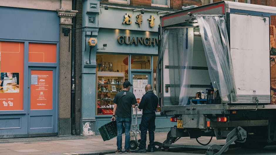 Two movers from Man with Van Knightsbridge are engaged in the loading process outside a shop on Sloane Street, Knightsbridge. They are dressed in dark clothing, with one wearing a cap, and are handling plastic crates, carrying them towards a large moving truck parked nearby. The truck features an open rear cargo area with plastic sheeting hanging from the sides, ready for furniture transport and packing during home relocation or local moving services. The shop's exterior has a blue facade with signs in Chinese characters and English, displaying various products through its large glass window. The surrounding pavement is clean, with the building's architectural details, such as decorative columns and a textured concrete wall, visible. This scene captures the logistics involved in house removals and furniture transport, emphasizing the careful handling of packing materials and equipment typical of professional removals services.