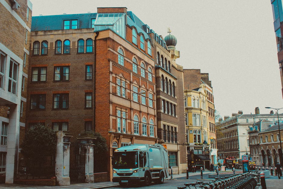 A daytime urban street scene showing the loading area for house removals in Sloane Street, Knightsbridge. Two large white removal vans are parked parallel to the pavement, one with the branding 'uniserve.co.uk' visible on the cabin, and the other with 'Bexton Transport' on its side. The vans are positioned close to a narrow storefront, with a motorcycle parked between them and the building on the left. The scene includes a pedestrian with a dark coat and backpack walking across the zebra crossing in front of the vans, and another individual with white hair and a dark coat walking on the opposite side of the crosswalk on the right. The surrounding buildings are classic stone-fronted structures with multiple windows, typical of a high-end London neighbourhood. The lighting indicates overcast weather, providing soft shadows and even illumination across the scene. This image exemplifies the logistical planning involved in furniture transport and home relocation in a busy city environment, as managed by professional moving services like Man with Van Knightsbridge.