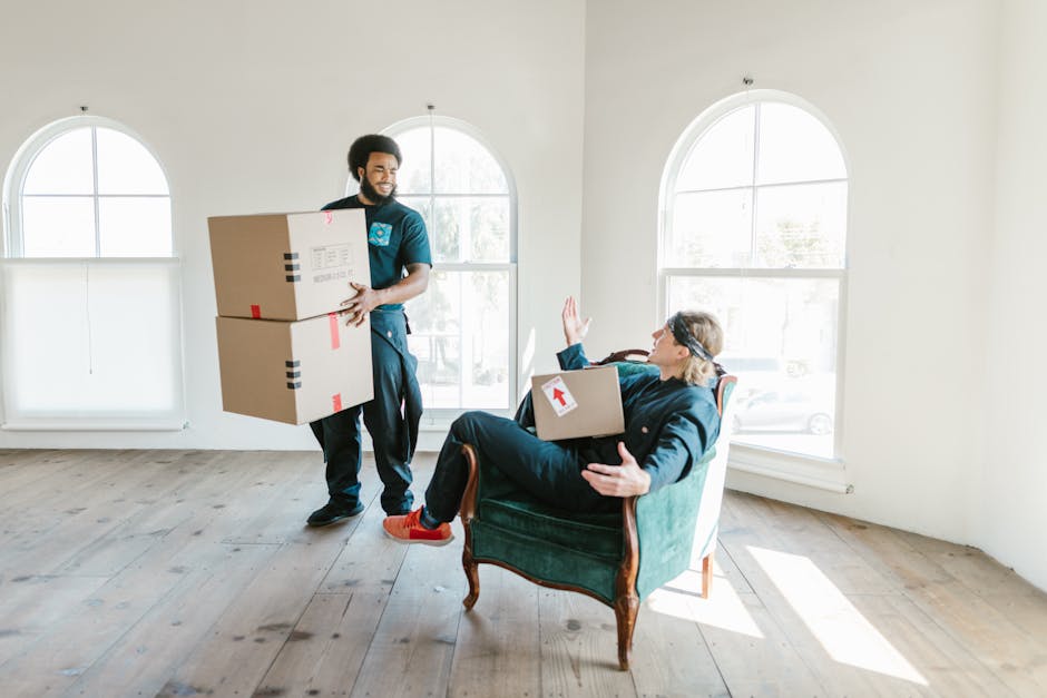 A man with dark hair and a beard, dressed in a dark blue uniform with a Man with Van Knightsbridge logo, is inside a bright, spacious room with white walls and large arched windows, carrying two medium-sized cardboard boxes with red arrow and tape markings indicating careful handling. He is approaching a woman seated in an antique green armchair, who is gesturing with her right hand and holding a similar box on her lap. The room has a light wooden floor and minimal furnishings, with sunlight streaming through the windows, suggesting a home environment preparing for a house removal. This scene captures the packing and loading process involved in home relocation, with professional movers handling boxes and furniture as part of a moving service.