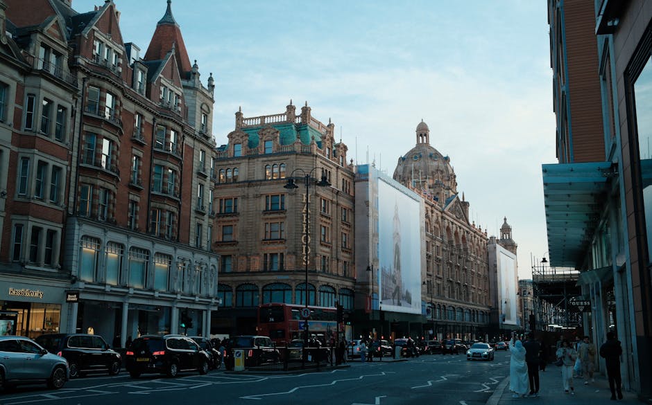 A busy street scene in Knightsbridge, London, with historic and modern buildings lining the pavement. Several parked cars and a row of mounted black street lamps are visible along the roadside. In the foreground, a group of people, some carrying luggage or moving boxes wrapped in plastic and cardboard, are preparing for home relocation or furniture transport. A red double-decker bus is approaching the curb, and a classic black taxi is also present. To the right, a building with glass storefronts and a café with outdoor seating suggests retail activity. The scene captures a typical urban environment where removals services by Man with Van Knightsbridge may operate, with a focus on loading and unloading furniture and boxes during packing and moving processes under daylight conditions.