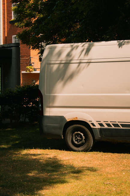 A white panel van is parked on a grassy area next to a sidewalk in a residential neighborhood, with a brick building and green trees in the background. The van's rear and side doors are closed, and the vehicle appears to be used for home relocation or furniture transport by Man with Van Knightsbridge. Shadows from the surrounding trees partially cover the van, and the scene suggests a quiet, sunny day suitable for loading or unloading furniture, boxes, and packing materials as part of a moving process. The van's black rubber bumper and wheel are visible, emphasizing its utility for professional removals services. This image represents the logistics involved in house removals within the Kensington and Chelsea area, highlighting the importance of parking rules for vans during a home move.