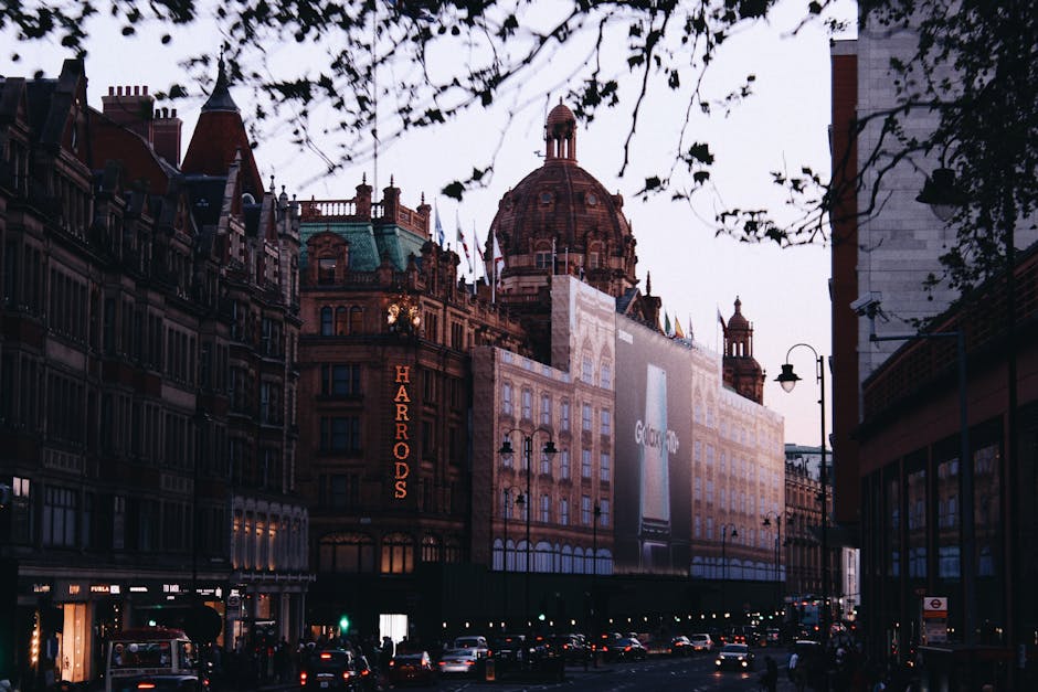 The image depicts a street view of Harrods department store in Knightsbridge, London, taken during the evening or late afternoon. The iconic building features ornate architecture with a prominent cent
