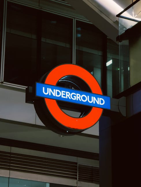 A close-up photograph of the iconic London Underground sign mounted on the exterior of a modern building at night. The sign features a red circular ring with a blue bar across the middle displaying the word 'UNDERGROUND' in white capital letters. The background includes glass windows and metal structures, illuminated by external lighting. The image captures the urban environment where house removals and furniture transport services, such as those provided by Man with Van Knightsbridge, often coordinate local moving logistics, including access restrictions at busy locations like Knightsbridge tube station for efficient packing, loading, and transportation of household items during home relocation projects.
