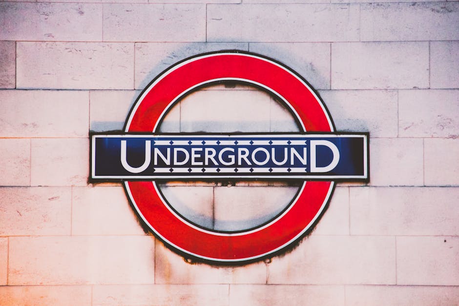 A close-up photograph of the London Underground roundel sign mounted on a light-colored brick wall. The sign features a red circle with a blue horizontal bar across the center, displaying the word 'UNDERGROUND' in white capital letters. The background setting suggests an indoor or outdoor location near the entrance or inside a property in Knightsbridge. This image emphasizes the station's branding, often relevant to home relocation or moving logistics involving transport routes near Knightsbridge tube station. Man with Van Knightsbridge provides professional removals and furniture transport services, and such signage can be relevant when coordinating pick-up or drop-off points within urban transport networks during packing and moving processes.