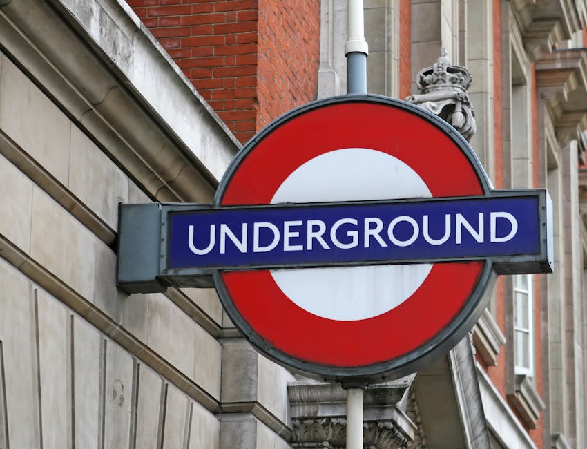 A close-up photograph of the iconic London Underground sign mounted on the exterior of a modern building at night. The sign features a red circular ring with a blue bar across the middle displaying the word 'UNDERGROUND' in white capital letters. The background includes glass windows and metal structures, illuminated by external lighting. The image captures the urban environment where house removals and furniture transport services, such as those provided by Man with Van Knightsbridge, often coordinate local moving logistics, including access restrictions at busy locations like Knightsbridge tube station for efficient packing, loading, and transportation of household items during home relocation projects.