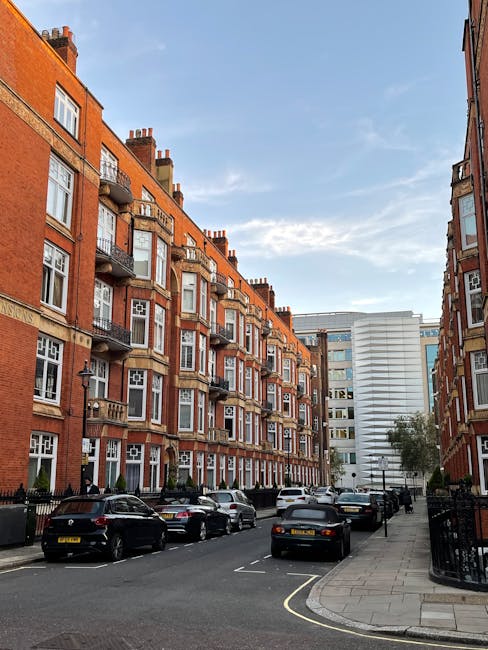 A street scene in Knightsbridge featuring a row of four to five-storey red-brick residential buildings with bay windows and small balconies, some of which have potted plants. Parked cars, including sedans and hatchbacks, line both sides of the narrow street. The pavement on the right side has a metal fence and some trees, while the sky above is partly cloudy with soft sunlight illuminating the buildings. Inside the property, visible through open doors or windows, there are cardboard boxes and wrapped furniture, indicating ongoing home relocation or packing activities, with some equipment like trolleys possibly being used for furniture transport. The overall scene suggests an urban environment undergoing a house removal or moving process, with [COMPANY_NAME] providing same-day removal services for urgent flat evacuations in Knightsbridge, as referenced on the page titled 'Same day Knightsbridge removals for urgent flat evacuations'.