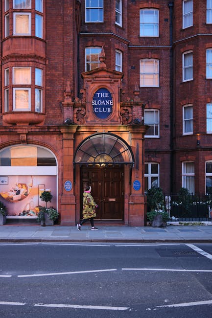 A woman dressed in a floral coat and black trousers is walking on the sidewalk in front of a multi-storey red brick building with large windows and a prominent arched entrance. Above the entrance, there is a blue sign that reads 'THE SLOANE CLUB,' and the doorway is framed by a semi-circular glass canopy. To the left of the entrance, a display window showcases interior furnishings, including a sofa and decorative plants, illuminated by warm lighting. The exterior features small potted plants and greenery near the base of the building, which is located in a London residential area. The scene takes place during daylight hours and captures the urban environment where professional house removals, such as those provided by Man with Van Knightsbridge, are often carried out. This setting reflects the typical context of moving logistics and furniture transport in tight access jobs, with the building entrance ready for loading or unloading during a home relocation or packing and moving process.