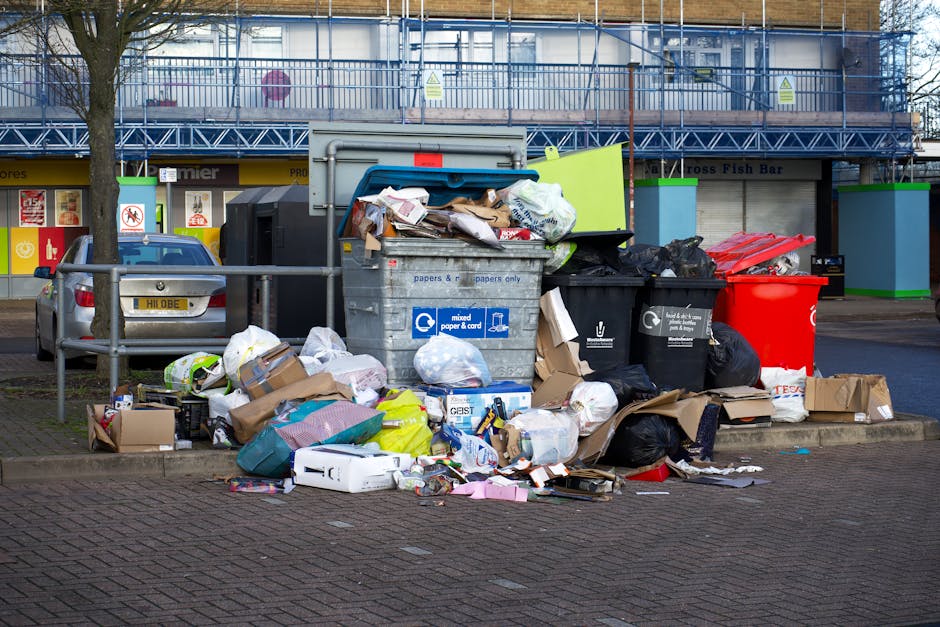 A collection of overflowing waste bins and scattered rubbish on a sidewalk, including large cardboard boxes, plastic bags filled with waste, paper, and packaging materials. The scene is set in front o
