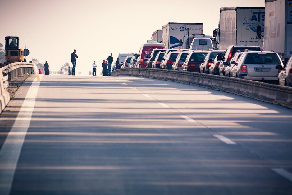 A traffic jam on a highway with multiple lanes of stationary vehicles including cars, delivery trucks, and larger freight trucks parked side by side. The scene is captured from a low angle, showing the vehicles on the right side and a barrier separating the lanes. Several people are visible on the pavement near the vehicles, some standing and others walking, possibly observing or waiting during the congestion. Shadows cast by the vehicles and objects indicate the time is either early morning or late afternoon with natural lighting. The scene reflects the challenges of heavy traffic and delays, relevant to planning for home relocation or furniture transport, as experienced during peak delays for Knightsbridge removals. Man with Van Knightsbridge, a professional removals service, would manage such logistics and ensure safe and efficient loading and transport of furniture and belongings amid traffic congestion, supporting clients with planning their moving schedules around peak times.