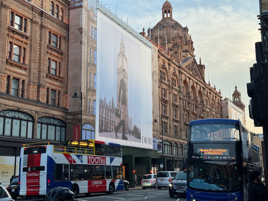 A busy street scene in Knightsbridge with historic building facades and a large Dior advertisement banner hanging between two structures. In the foreground, a double-decker bus with a digital route display and a red-and-white tour bus are visible, both paused in traffic. Several cars are also waiting at the intersection, with some passengers visible inside the vehicles. Pedestrians are walking along the pavement and waiting at the roadside, some wearing face masks. The scene is illuminated by natural daylight, with the sky partly cloudy. The architectural style of the buildings features ornate details, domes, and spires, characteristic of classic London architecture. This image illustrates typical urban surroundings involved in home relocations or furniture transport activities, often requiring logistics coordination by services like Man with Van Knightsbridge, especially during peak delays for house removals in this area.